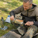 Man eating Trek'N Eat Spicy Beef Casserole freeze dried camping meal outdoors on grass with stove and gear nearby