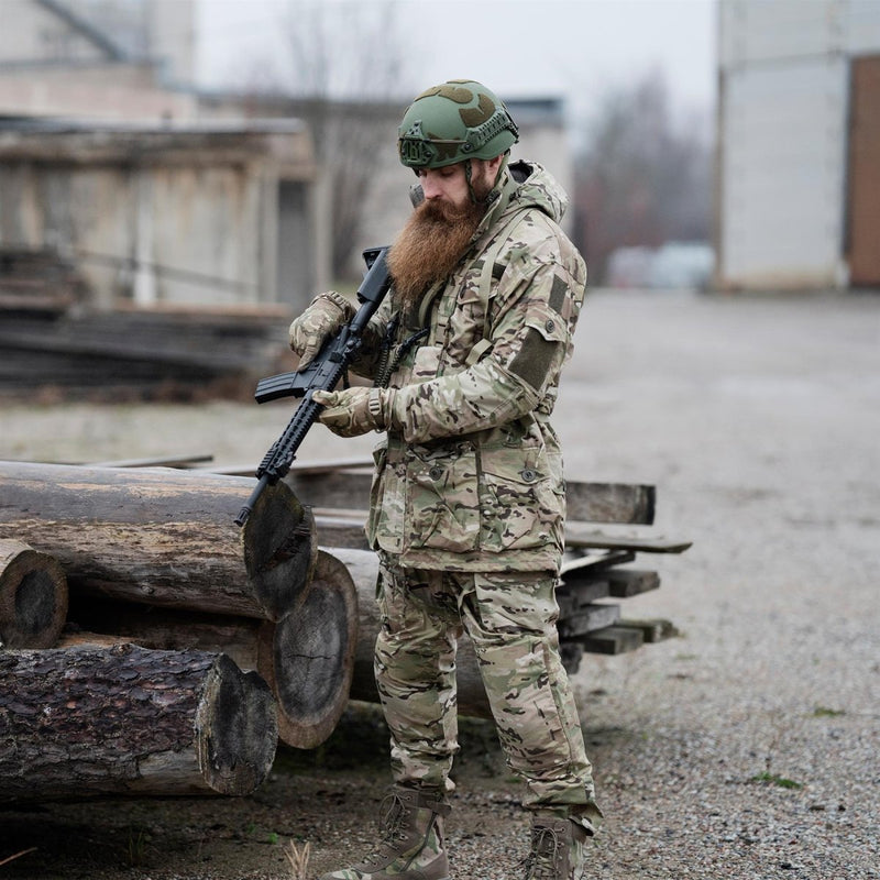 Man wearing Leo Kohler tactical field pants in multicam camouflage holding a rifle outdoors near logs