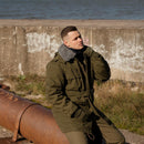Man in a green jacket sitting on a rusted pipe by a concrete wall and water.